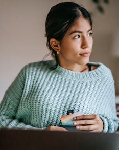 Young dreamy ethnic female buyer with debit card and netbook looking away in house room